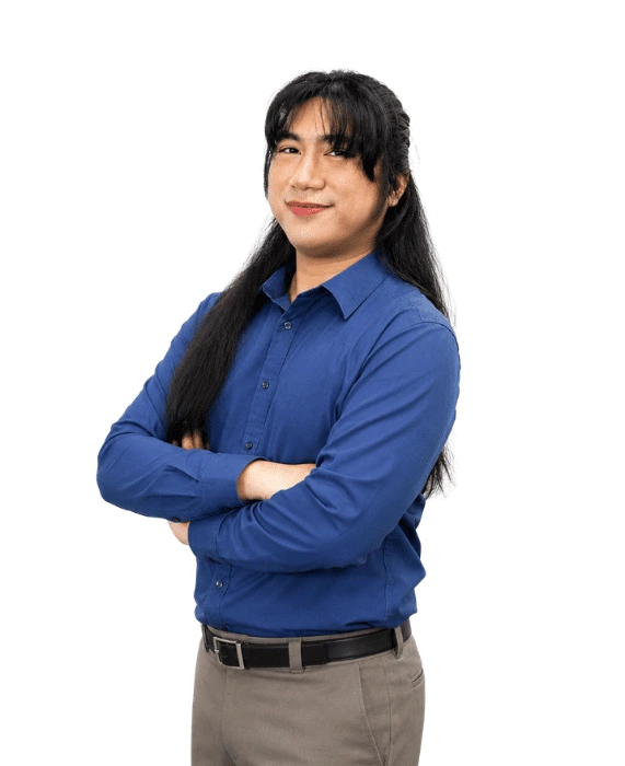 Professional portrait of a person wearing a blue long-sleeve button-down shirt and gray trousers, standing with arms crossed and smiling confidently against a light background.